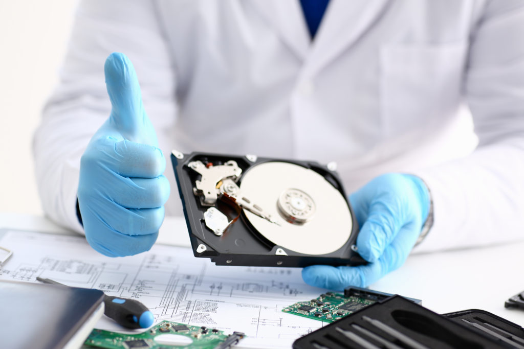 A technician wearing blue gloves while holding a hard drive from computer laptop showing a thumbs up gesture indicating that the data recovery is successful 