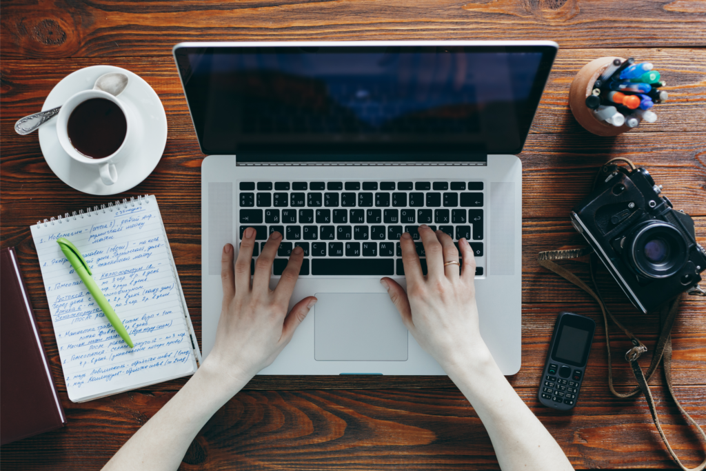 A person working on a computer with a pen and paper, camera, phone, and coffee on both sides 