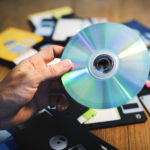 A person holding a CD ROM with a lot of floppy disks in the background